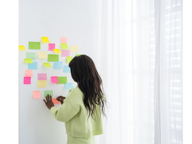 A woman in a light green outfit arranging colorful sticky notes on a white wall, planning ideas in a bright, modern workspace.