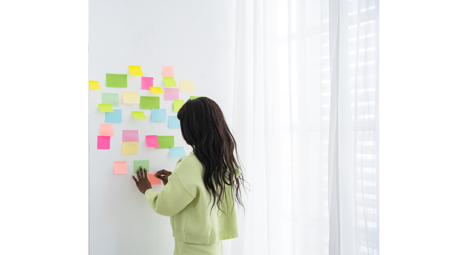 A woman in a light green outfit arranging colorful sticky notes on a white wall, planning ideas in a bright, modern workspace.