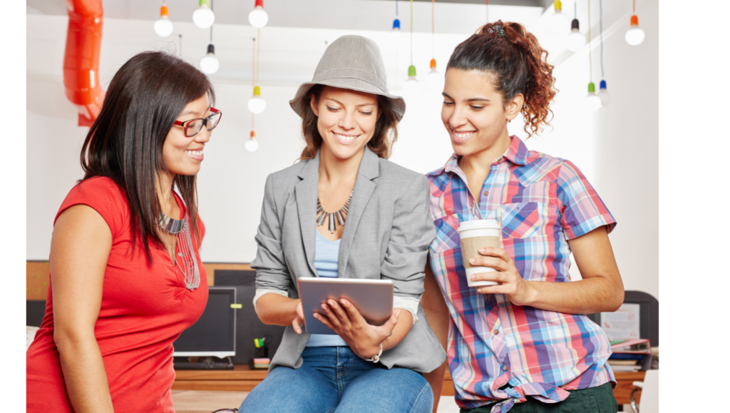 Three women in a creative workspace looking at a tablet, discussing ideas while enjoying coffee and smiling.