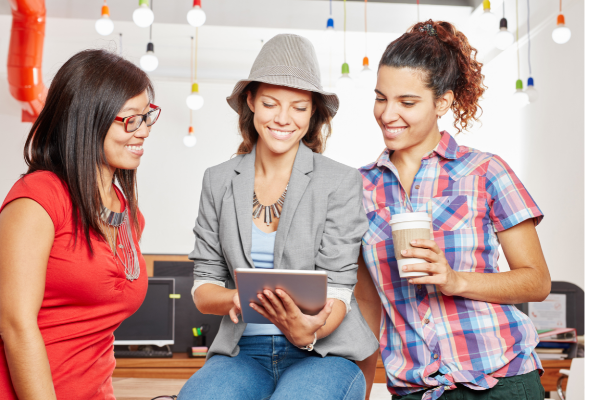 Three women in a creative workspace looking at a tablet, discussing ideas while enjoying coffee and smiling.