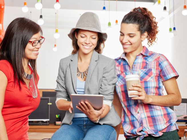 Three women in a creative workspace looking at a tablet, discussing ideas while enjoying coffee and smiling.